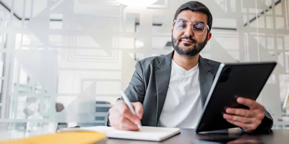 Professioneller Geschäftsmann mit Brille arbeitet an einem Tablet und macht sich Notizen in einem hellen, modernen Büro