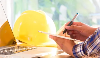 Mitarbeiter arbeitet am Schreibtisch im Homeoffice, neben dem Laptop liegt ein Schutzhelm. © Shutterstock, oekka.k