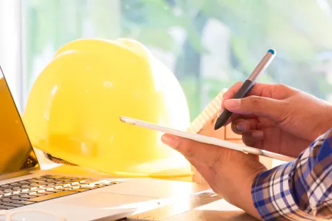 Mitarbeiter arbeitet am Schreibtisch im Homeoffice, neben dem Laptop liegt ein Schutzhelm. © Shutterstock, oekka.k