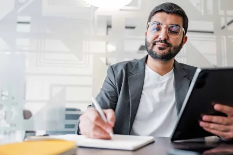 Professioneller Geschäftsmann mit Brille arbeitet an einem Tablet und macht sich Notizen in einem hellen, modernen Büro