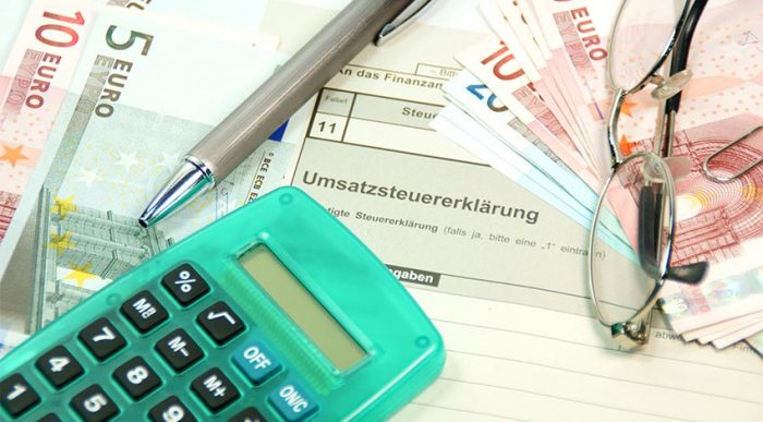 Ein Taschenrechner, ein Stift und eine Brille auf einem Stapel Papier und Euroscheinen hinsichtlich der Umsatzsteuererklärung. Die Umsatzsteuergrenze wurde durch das Wachstumschancengesetz angehoben. © Adobe Stock, PhotographyByMK