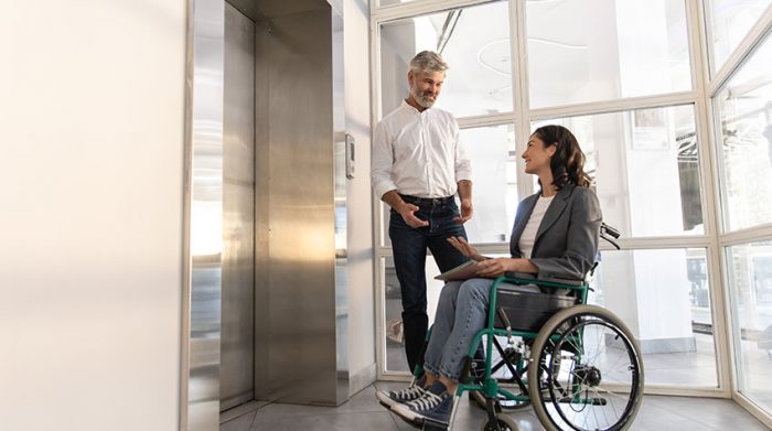 Mann spricht mit lächelnder Frau im Rollstuhl vor einem Aufzug in einem modernen Bürogebäude.